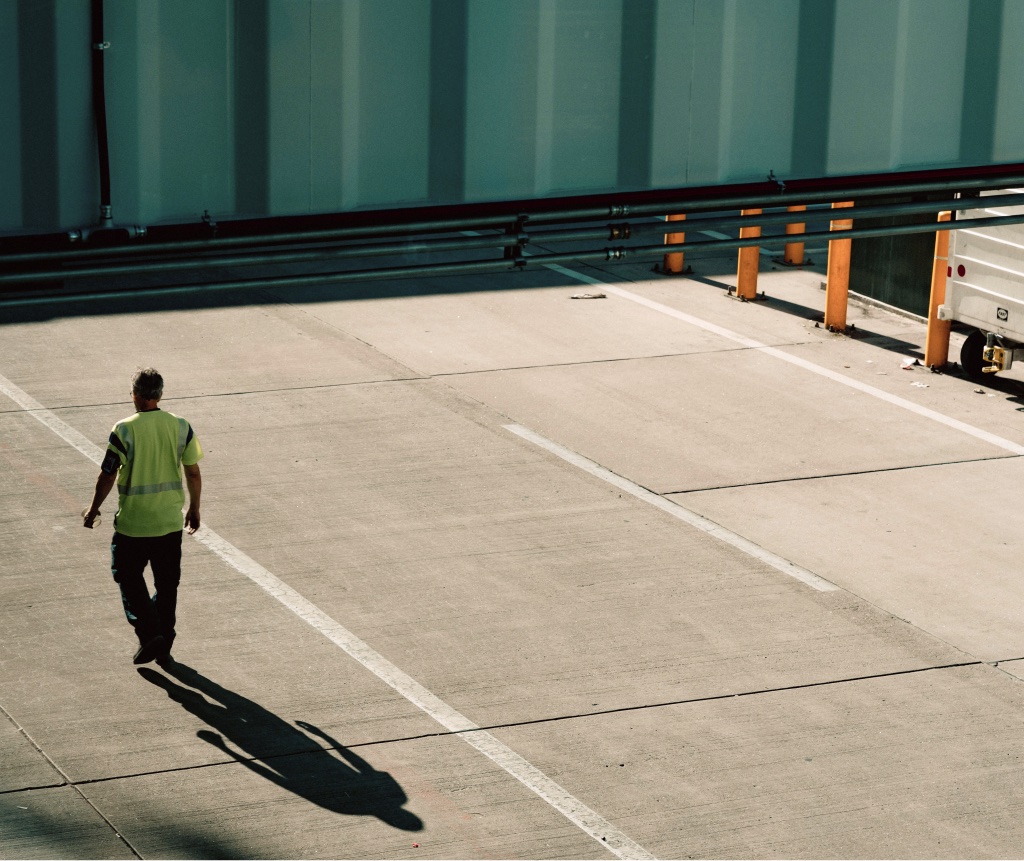 A worker in a high-visibility vest walking across a sunlit loading yard, his long shadow behind him